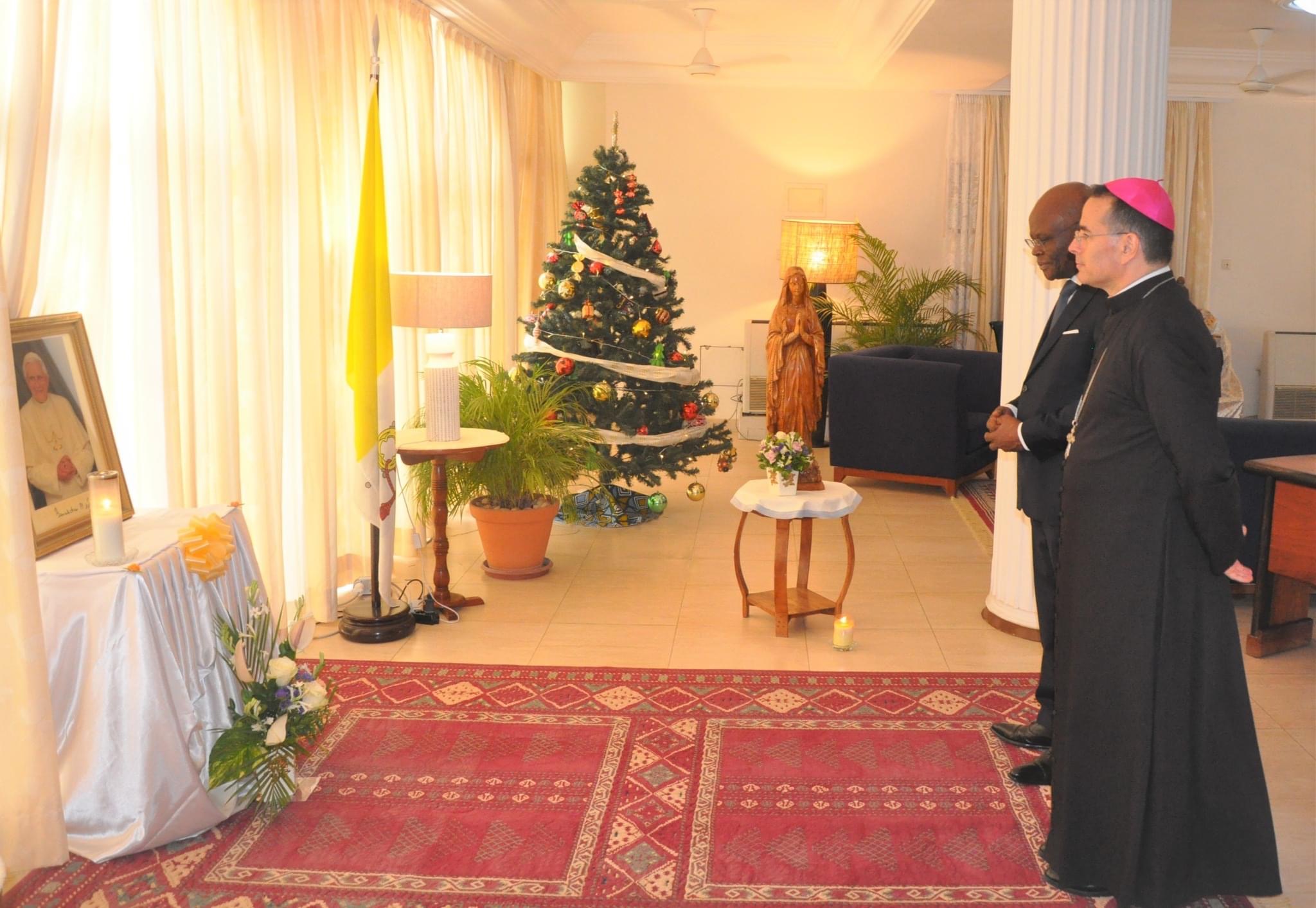 Deux hommes en habits religieux et formels regardent un portrait posé sur une table entourée de fleurs et bougies dans une pièce décorée pour Noël