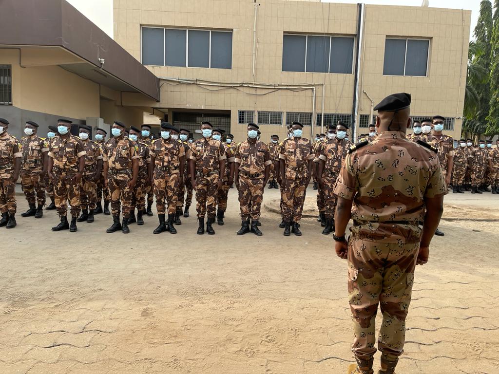 Un groupe de militaires en tenue camouflage alignés à l'extérieur face à un homme debout dos à la caméra.