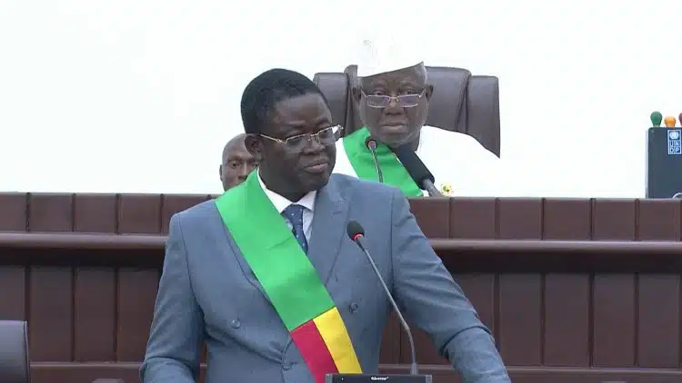 Homme en costume gris et écharpe verte, jaune et rouge parle au micro dans une salle officielle avec deux hommes assis derrière lui