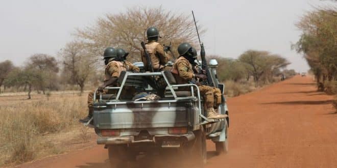 Quatre soldats en tenue et casque sont armés dans la benne d un pick-up sur une piste en terre rouge bordée d arbres secs