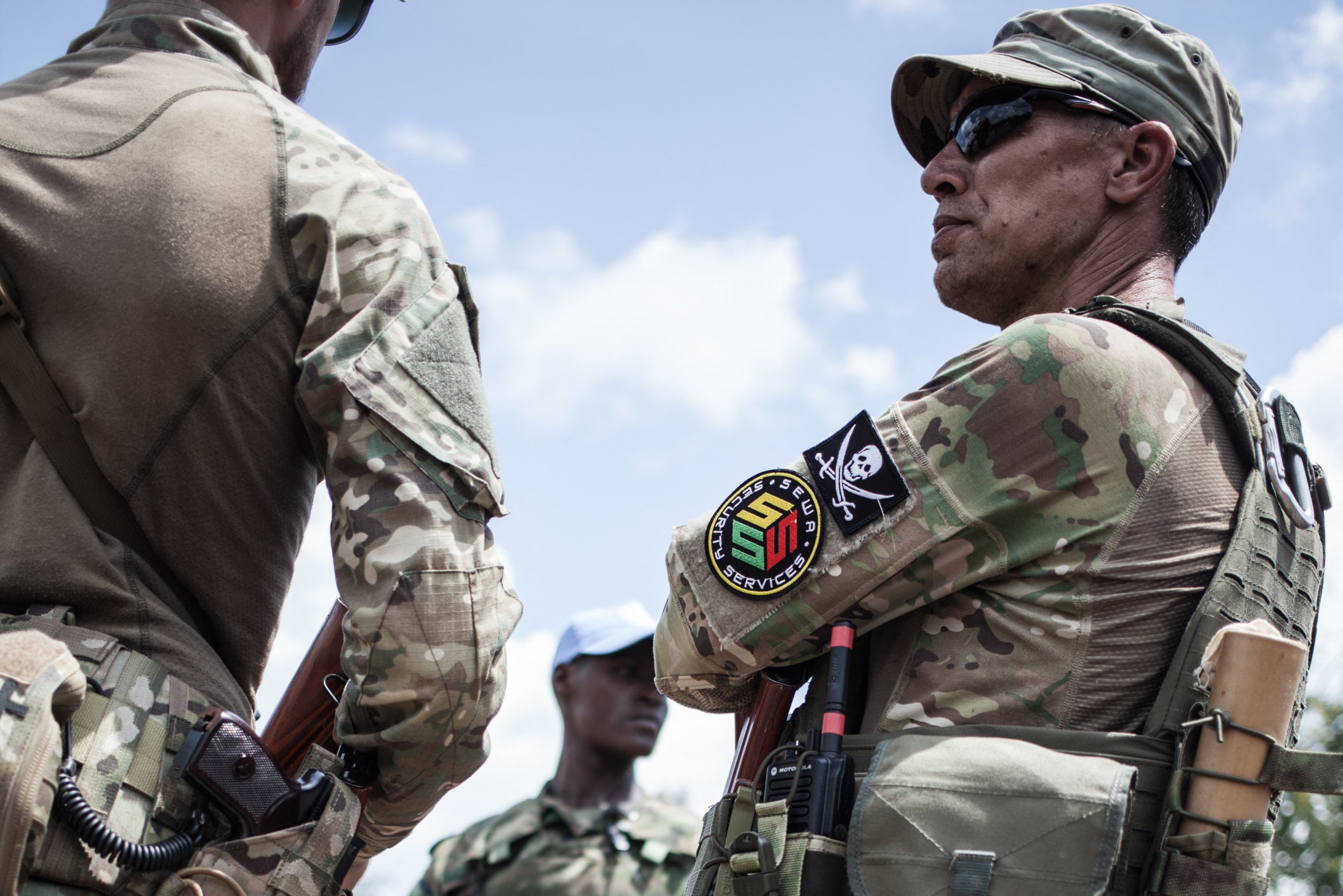 Deux hommes en tenue militaire camouflage discutent avec un troisième en arrière-plan au ciel bleu partiellement nuageux