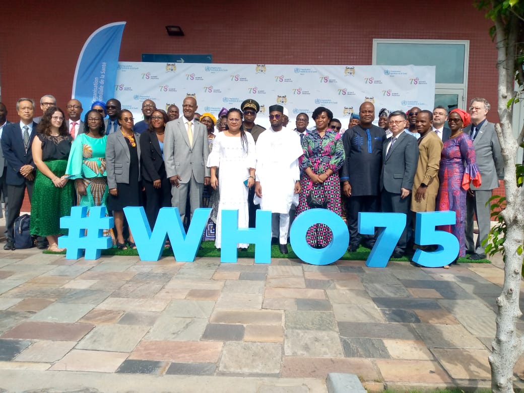 Groupe de personnes posant debout derrière un grand panneau bleu #WHO75 devant un mur décoré de slogans célébrant 75 ans