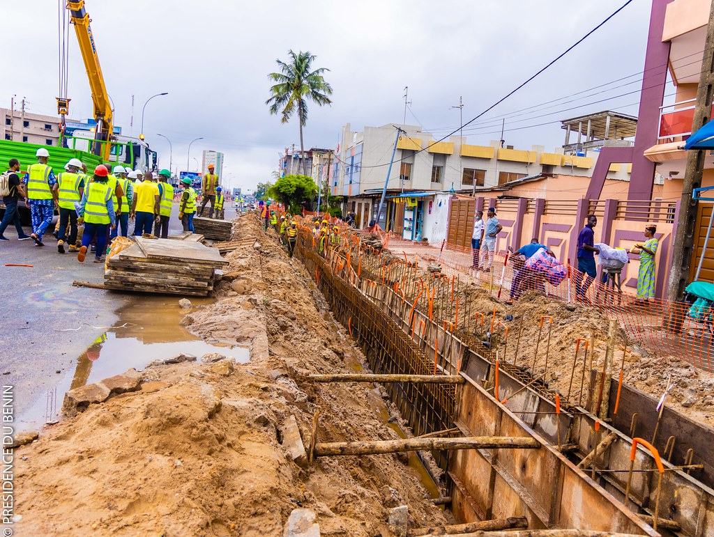Un chantier de construction à ciel ouvert en milieu urbain avec des ouvriers et passants le long d une tranchée profonde.
