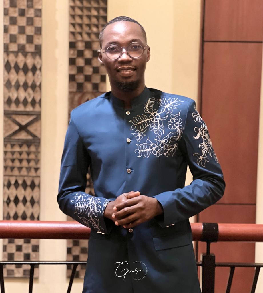 Homme debout en tenue traditionnelle bleu avec motifs blancs, lunettes rondes, mains jointes devant un décor intérieur.