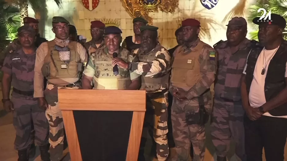 Un groupe d’hommes en tenue militaire se tient derrière un pupitre avec un microphone dans une pièce ornée de blasons muraux