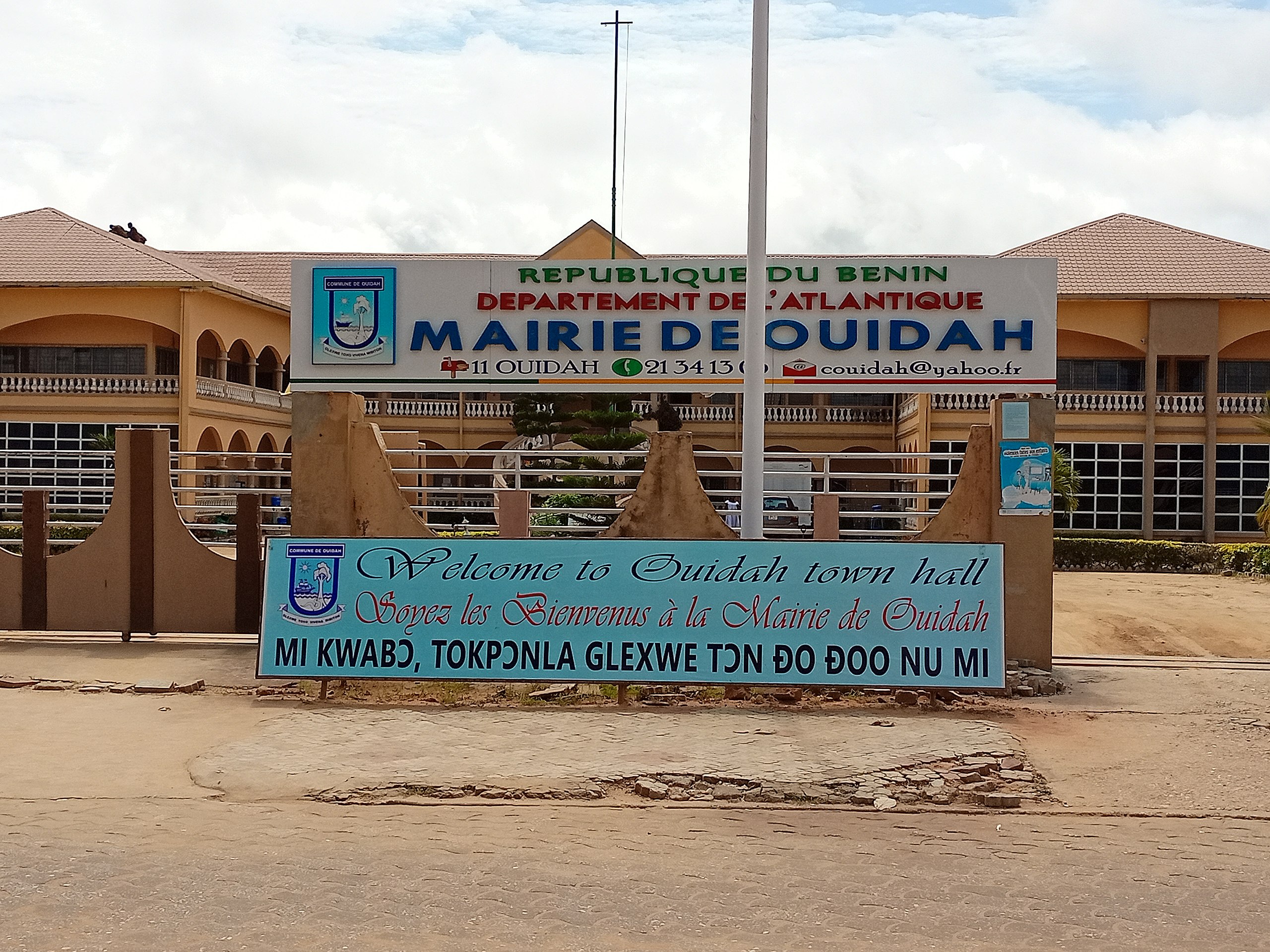 Façade d un bâtiment municipal avec plusieurs panneaux indiquant la mairie de Ouidah et un message de bienvenue multilingue