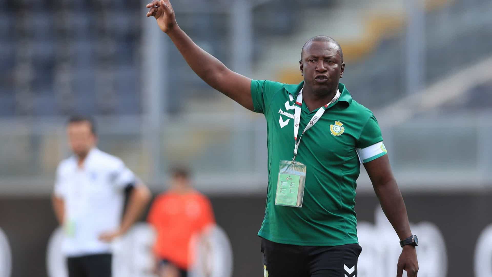 Un homme en polo vert donne une instruction avec le bras levé dans un stade flou en arrière-plan