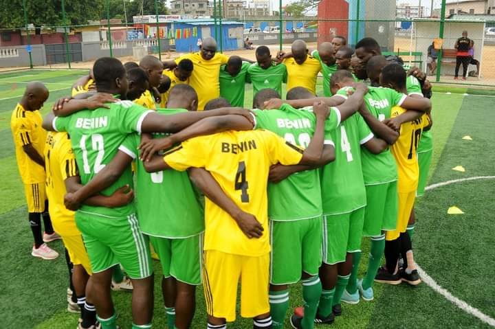 Joueurs de football en cercle sur un terrain artificiel en tenues jaunes et vertes avec inscriptions Benin sur les maillots