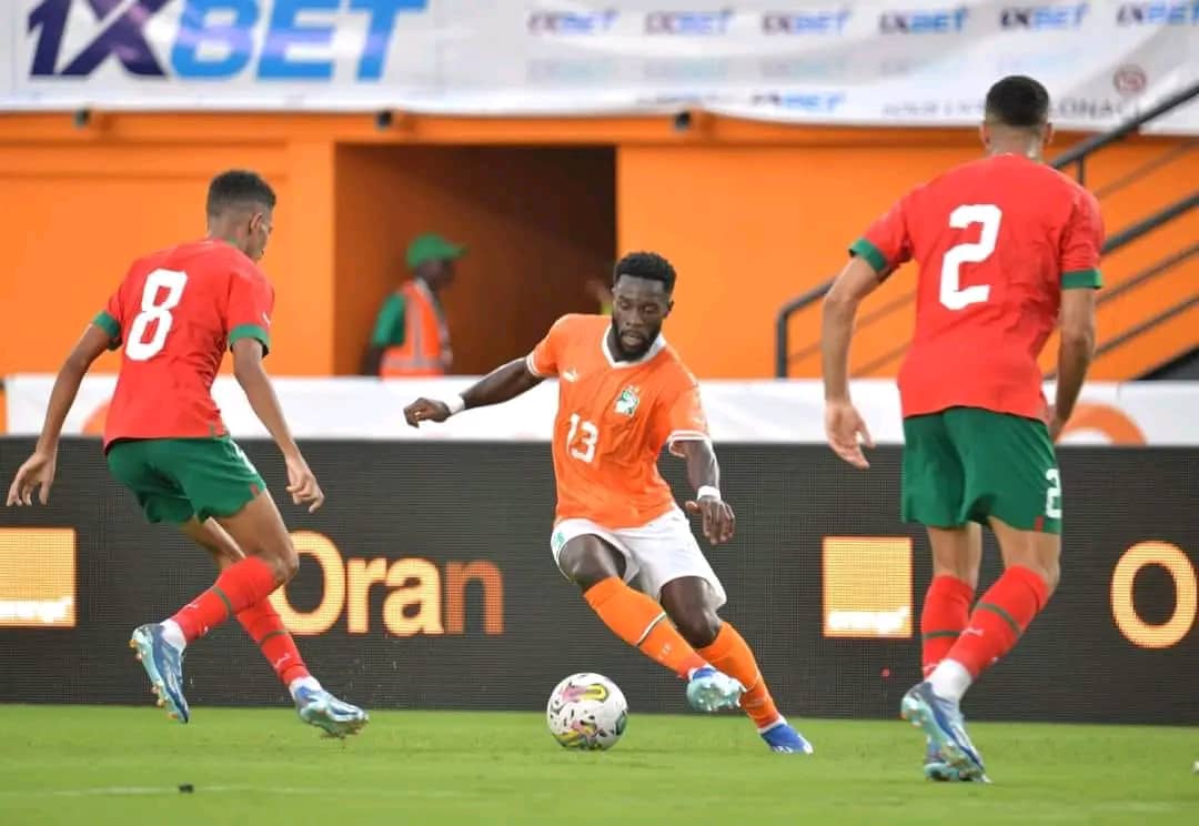 Un joueur en maillot orange contrôle le ballon face à deux joueurs en maillot rouge et vert sur un terrain de football