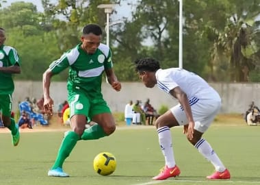 Deux joueurs de football en action sur un terrain gazonné devant des spectateurs assis au bord du terrain