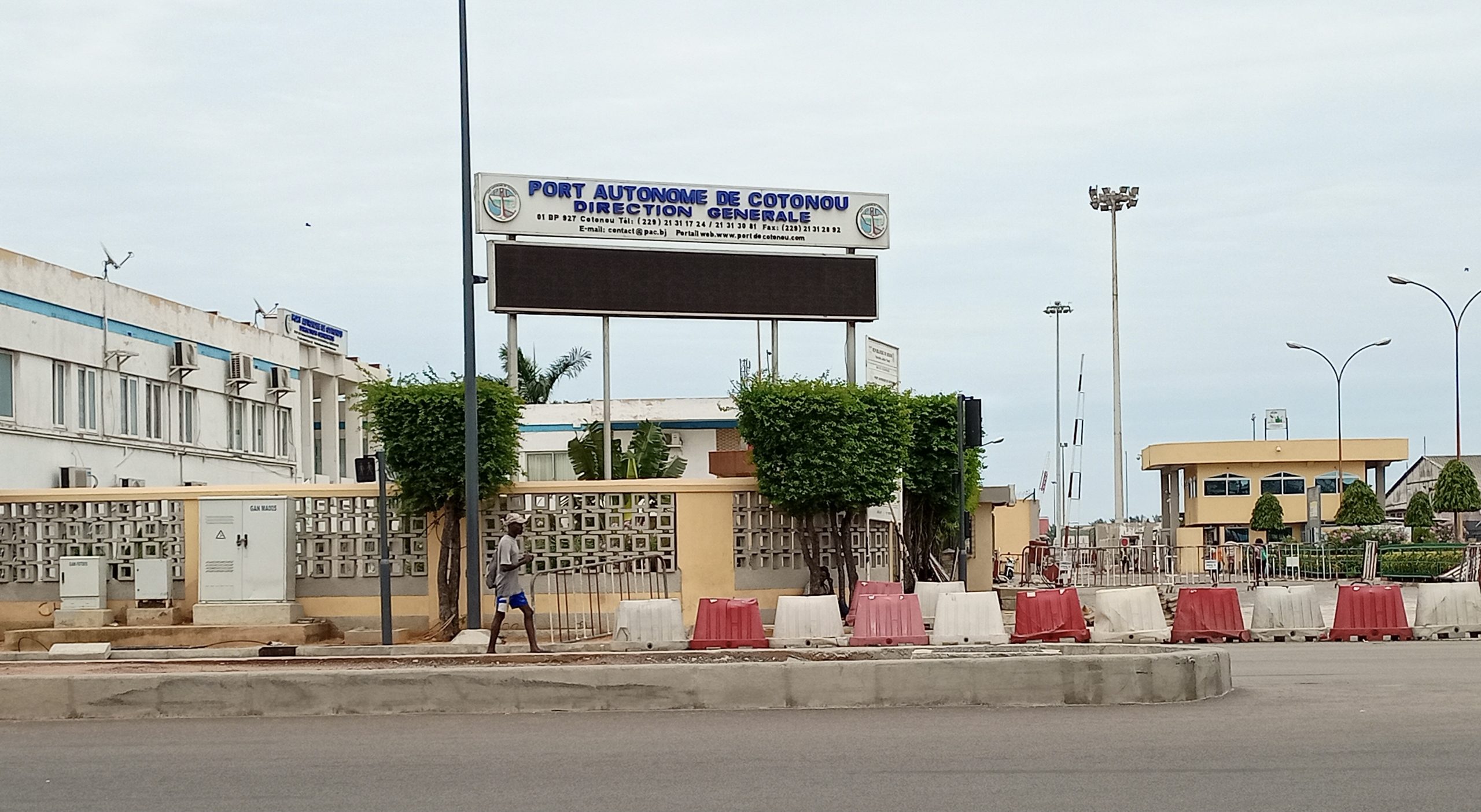 Entrée d'un bâtiment administratif avec un panneau Port Autonome de Cotonou Direction Générale et des barrières de chantier rouges et blanches.