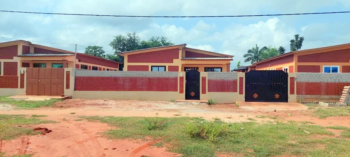 Maisons mitoyennes avec murs en briques rouges et portails métalliques noirs dans un quartier en terre battue et herbes clairsemées
