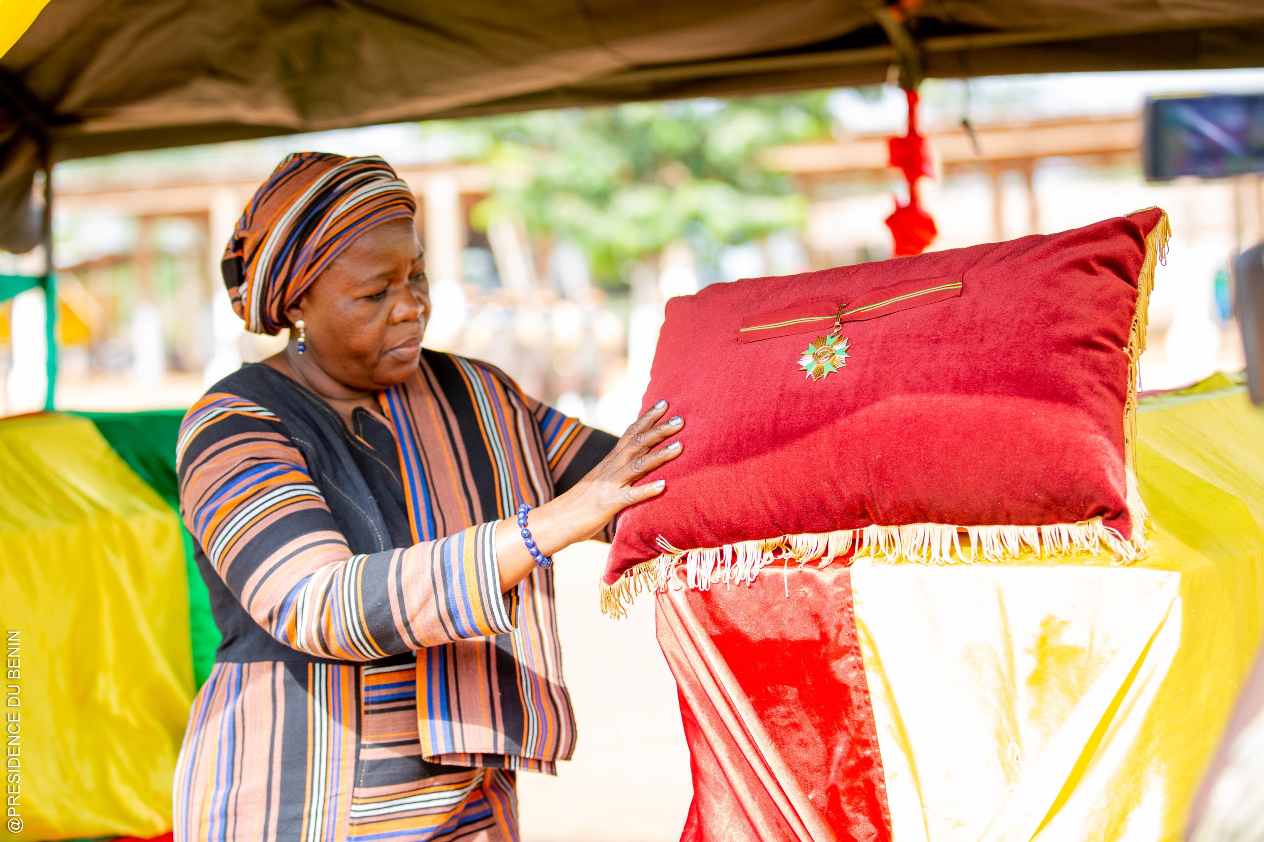 Femme en tenue rayée touchant un coussin rouge orné d'une décoration militaire sous un abri en extérieur