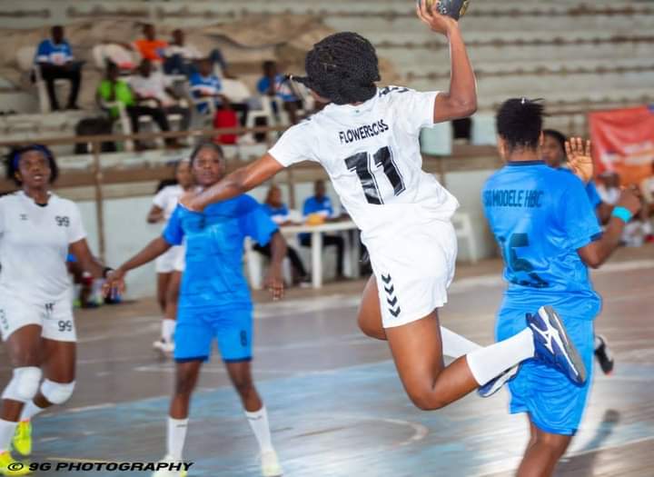Une joueuse en blanc saute pour lancer le ballon face à des adversaires en bleu lors d'un match de handball en salle
