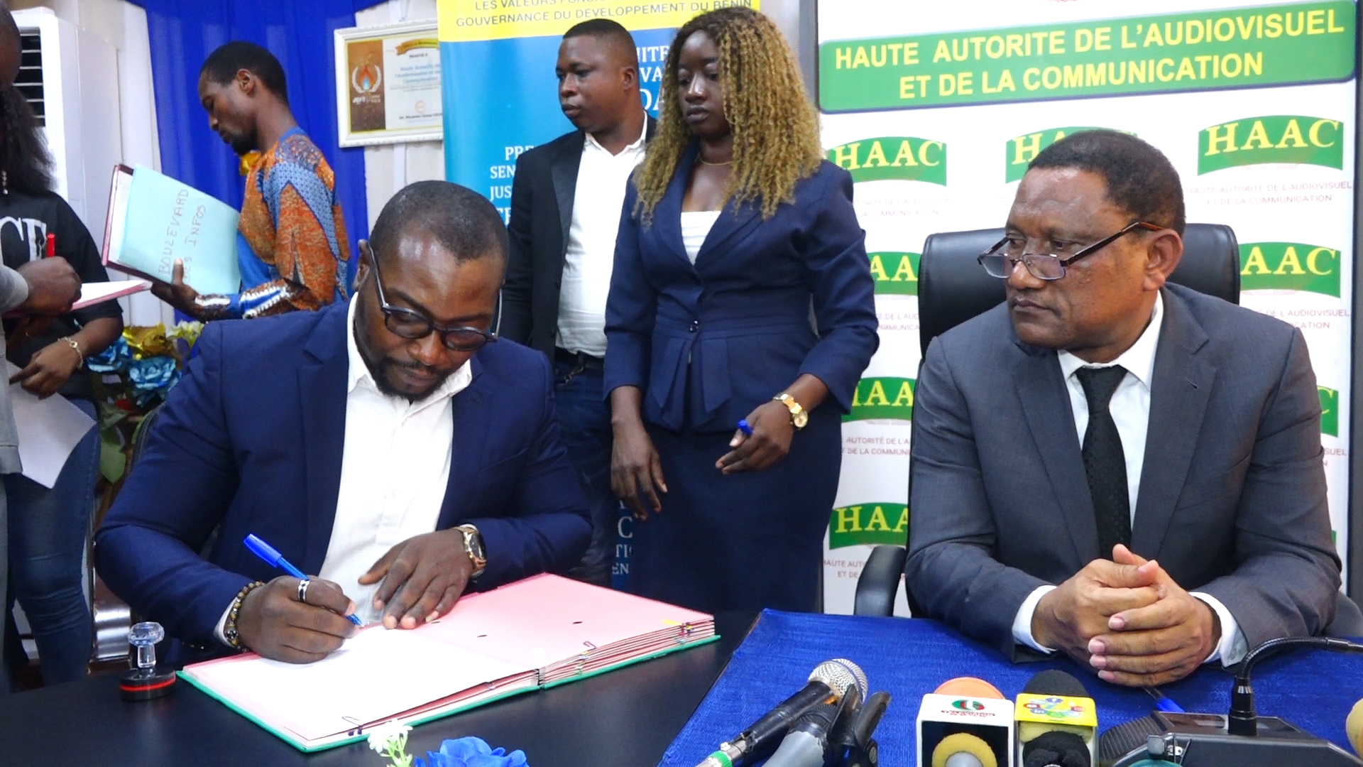 Un homme signe un document à une table tandis qu'un autre homme en costume sombre et trois personnes posent en arrière-plan devant des panneaux HAAC