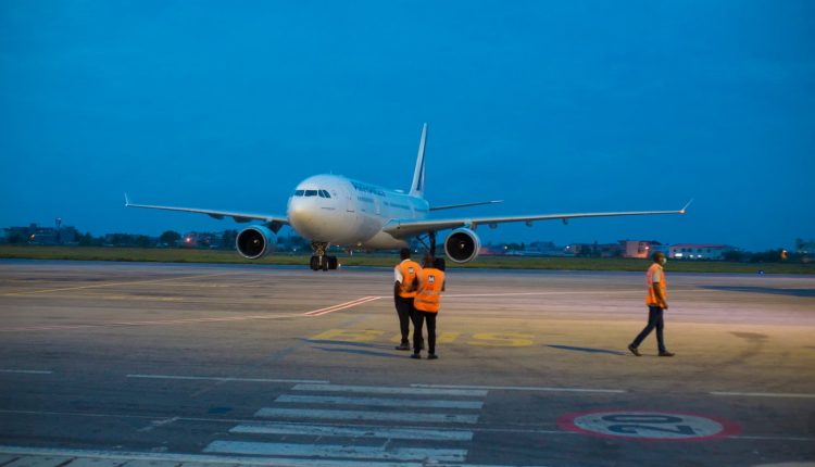 Avion stationné sur la piste d’aéroport avec trois personnels en gilets orange au premier plan au crépuscule