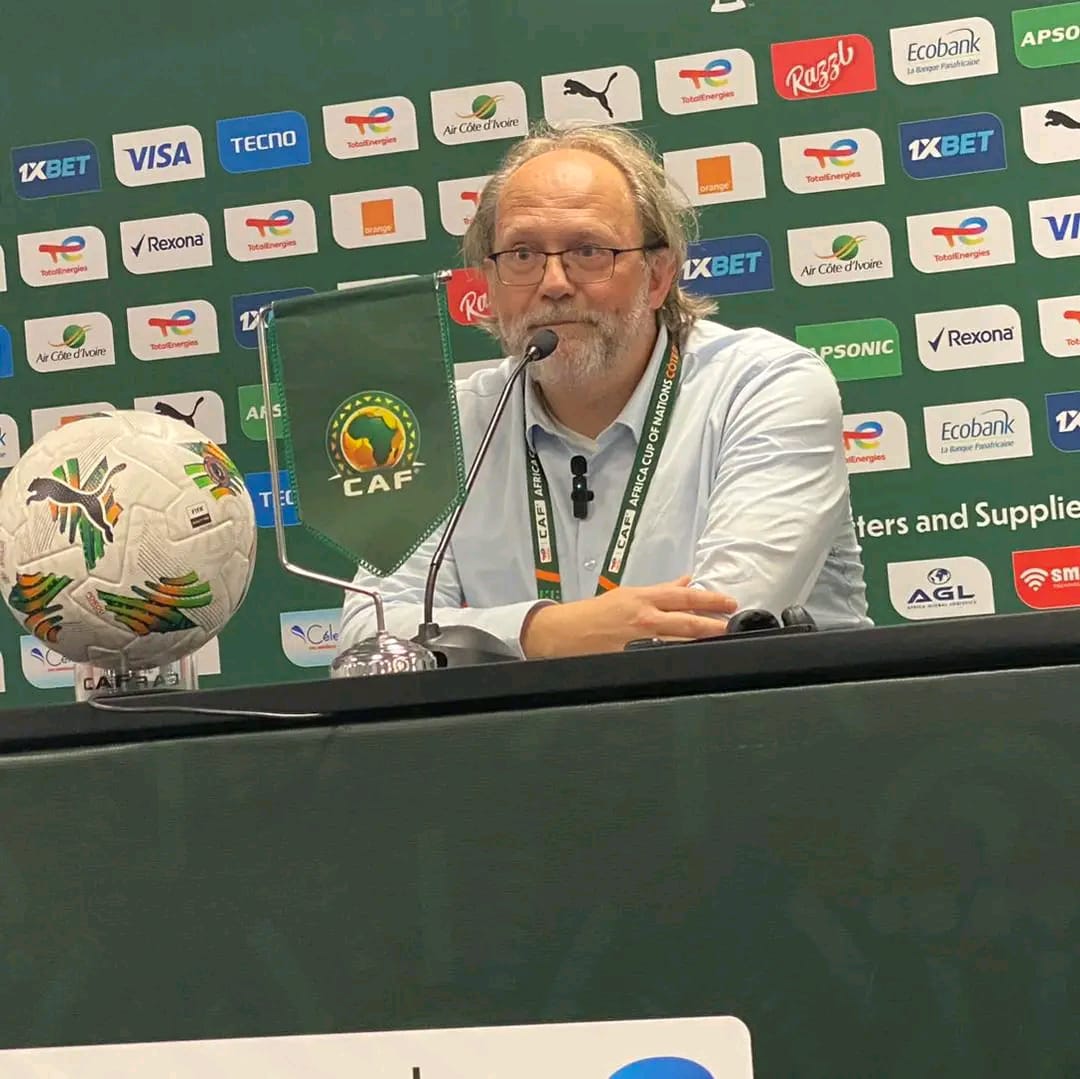 Un homme portant des lunettes est assis à une table avec un micro devant un fond marqué de divers logos sportifs et un ballon de football posé sur la table