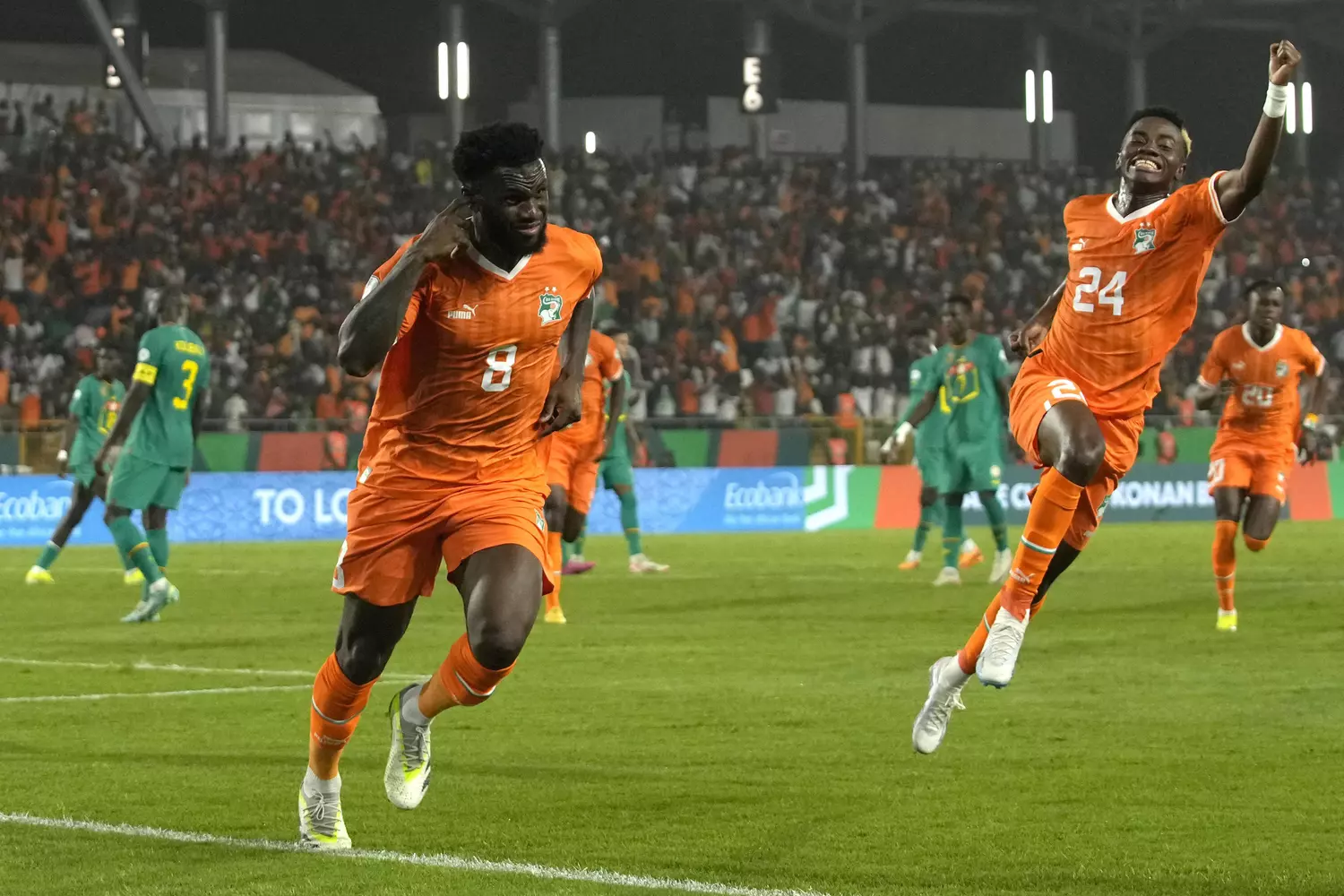 Deux joueurs en maillot orange célèbrent un but sur un terrain de football face à une équipe en vert devant un stade plein