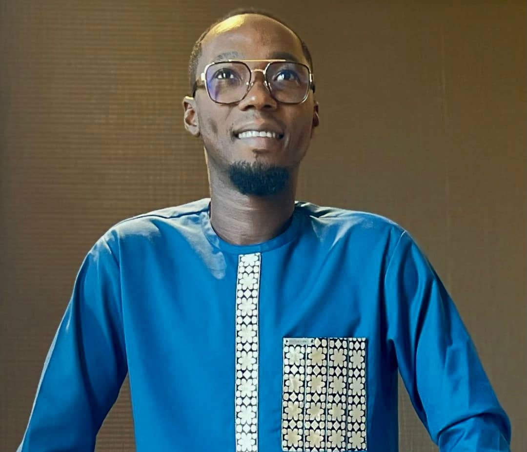 Homme souriant portant une chemise bleue avec un motif floral clair sur le devant et de grandes lunettes dans un intérieur aux murs marron