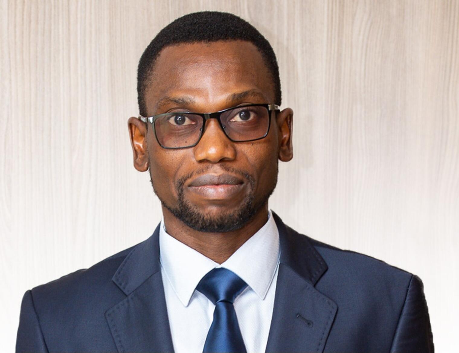 Homme portant des lunettes habillé en costume bleu, cravate et chemise blanche, regardant devant lui sur fond clair