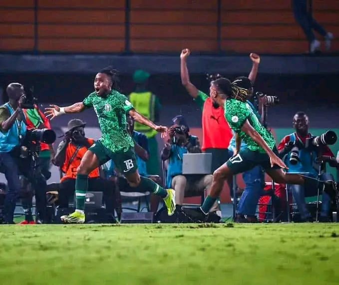 Deux joueurs en maillot vert célèbrent en courant sur un terrain de football devant des photographes assis au bord de la pelouse