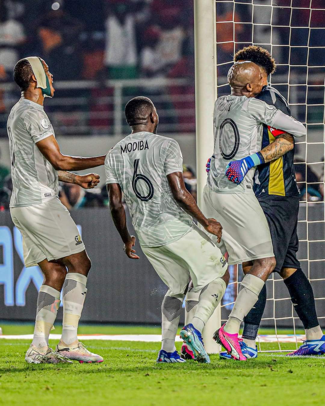 Quatre joueurs de football en tenue claire célèbrent ensemble près du but sur un terrain en soirée