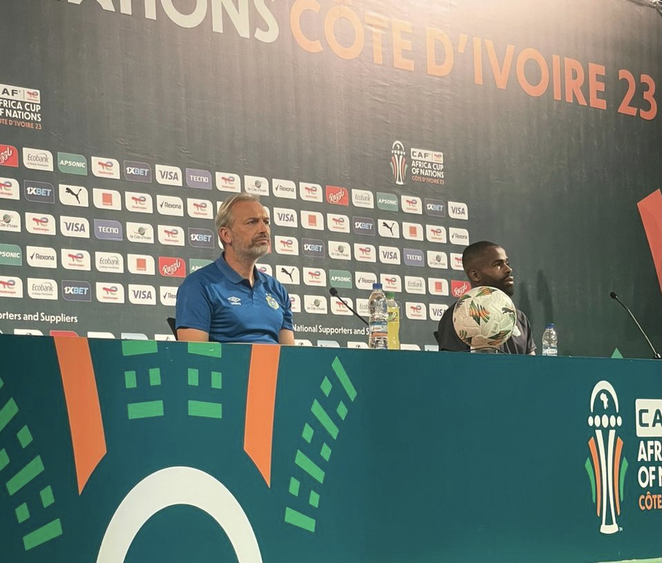 Deux hommes assis à une table de conférence avec un ballon de football et un fond décoré pour une compétition sportive africaine.
