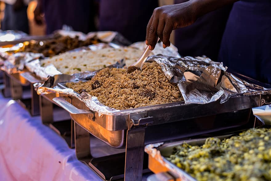 Main en train de servir du riz dans un plat en inox lors d'un buffet avec plusieurs plats disposés côte à côte