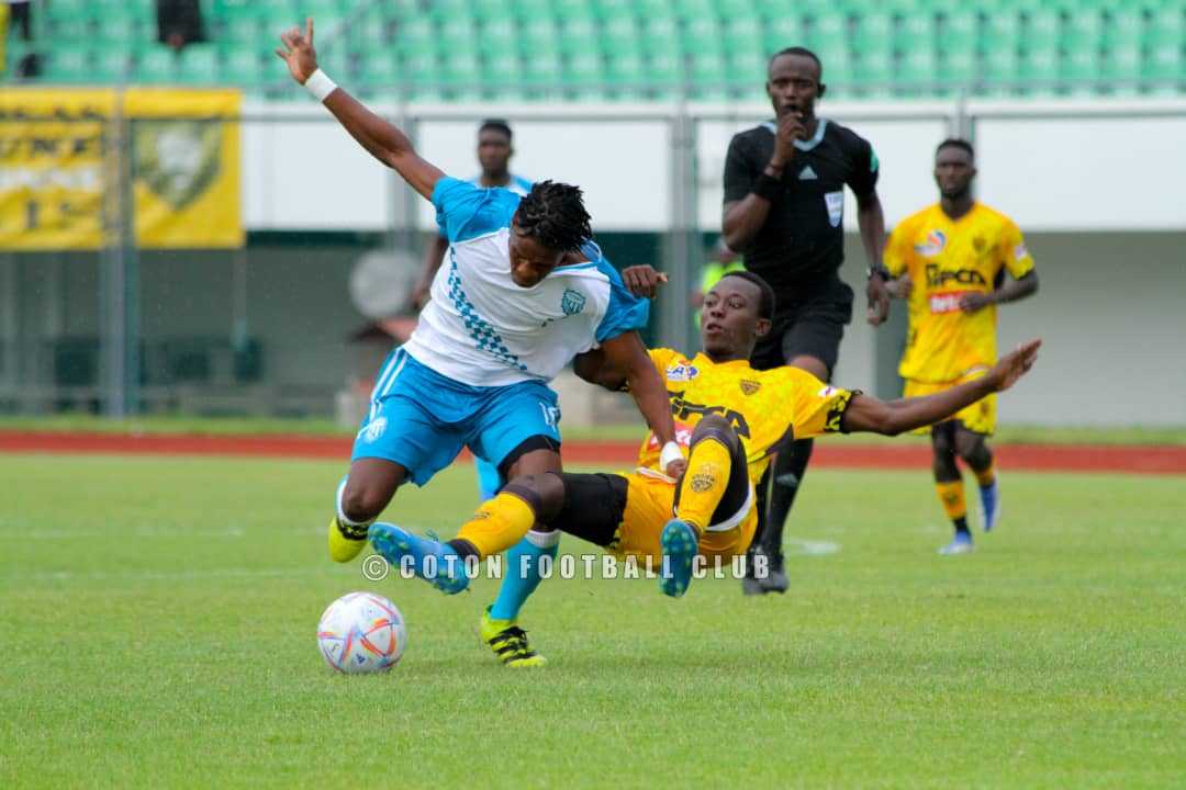 Un joueur en maillot bleu chute en tentant de dribbler un défenseur en jaune sur terrain vert devant un arbitre en noir et d'autres joueurs.