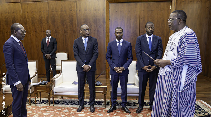 Cinq hommes en costume et un homme en tenue traditionnelle debout dans une salle avec des fauteuils et un tapis rouge