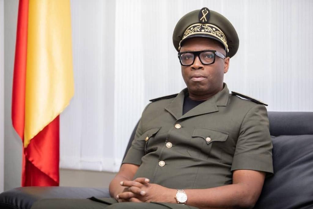 Homme en uniforme militaire assis sur un canapé avec un drapeau rouge et jaune en arrière-plan
