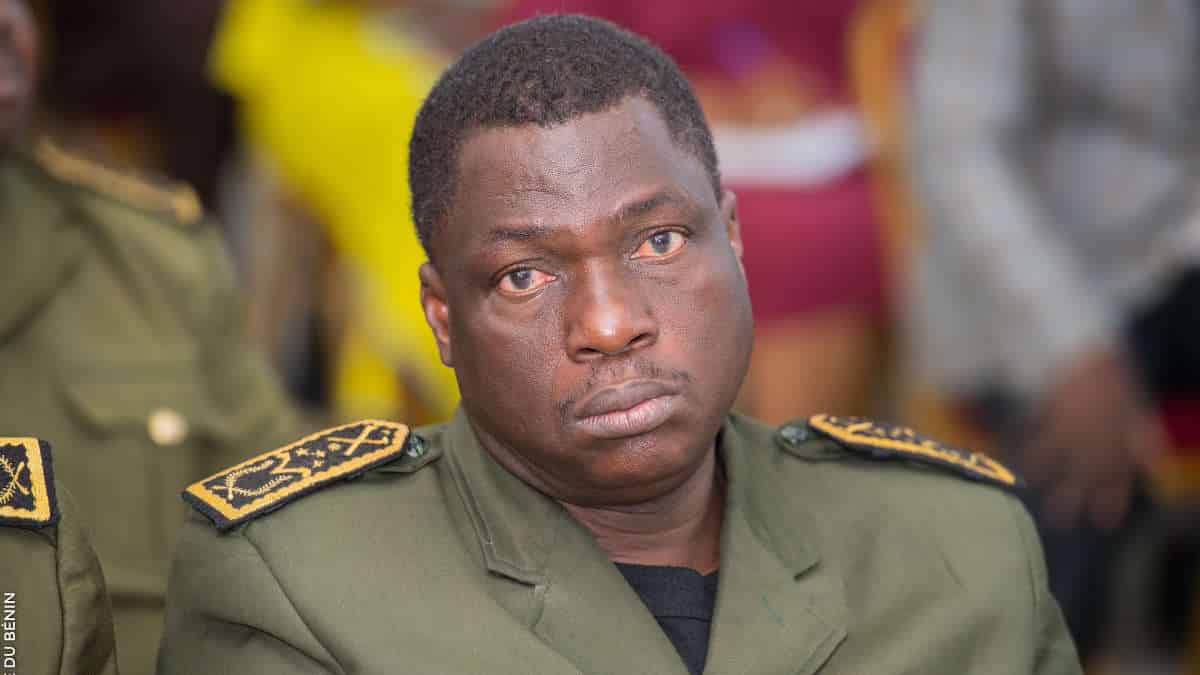 Homme en uniforme militaire vert regardant vers l'avant avec une expression sérieuse en arrière-plan flou coloré