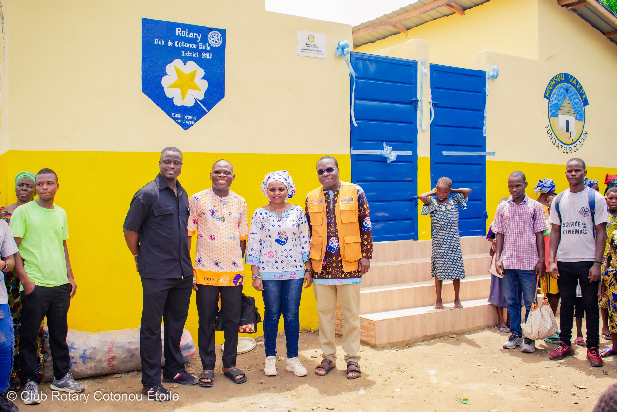 Un groupe de personnes posant devant un bâtiment jaune avec des portes bleues et des panneaux sur le mur.