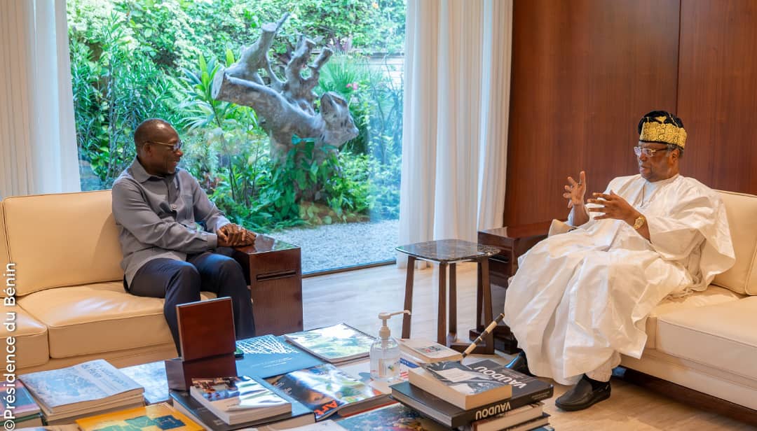 Deux hommes discutent assis dans un salon où un grand canapé clair, une table basse avec livres et une sculpture sont visibles