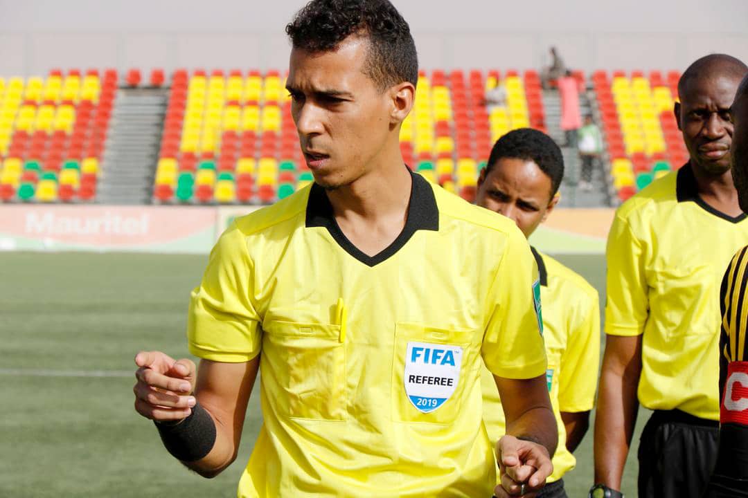 Un arbitre en maillot jaune avec un badge FIFA marche sur un terrain de football devant des gradins colorés rouge et jaune