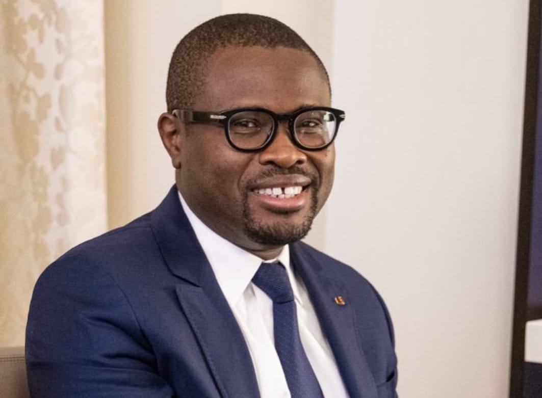 Homme en costume bleu, chemise blanche et cravate bleue souriant et portant des lunettes noires assis dans un intérieur clair