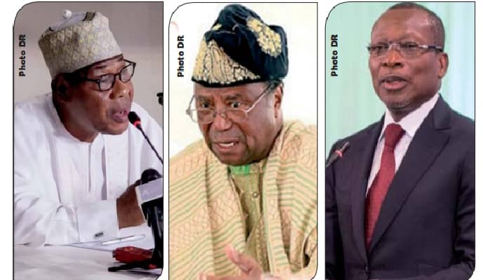 Trois hommes en gros plan, l’un en tenue traditionnelle blanche parlant devant un micro, le deuxième en pagne et bonnet traditionnels, le troisième en costume-c