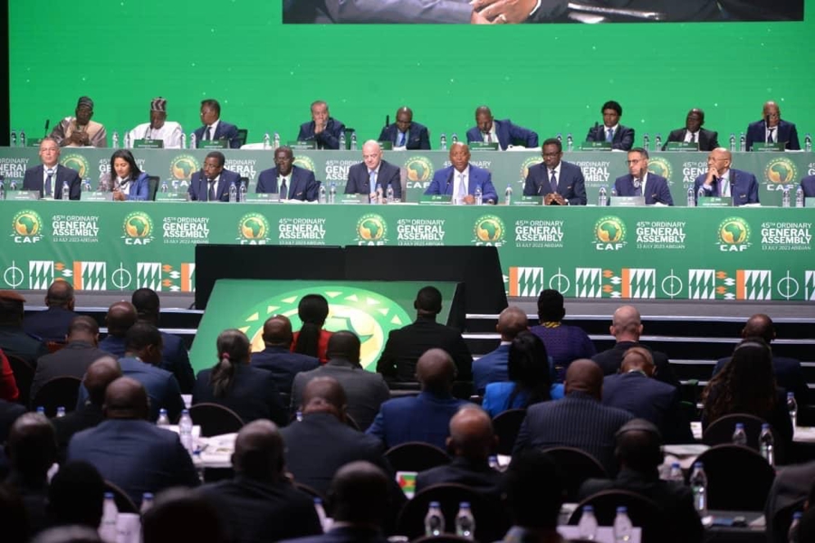 Personnes assises en rangées face à une scène avec un grand écran vert et logos CAF General Assembly