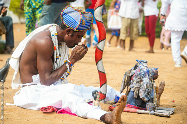 Homme en tenue traditionnelle assis par terre devant un petit autel orné de statuettes et de tissus colorés dans un espace en plein air