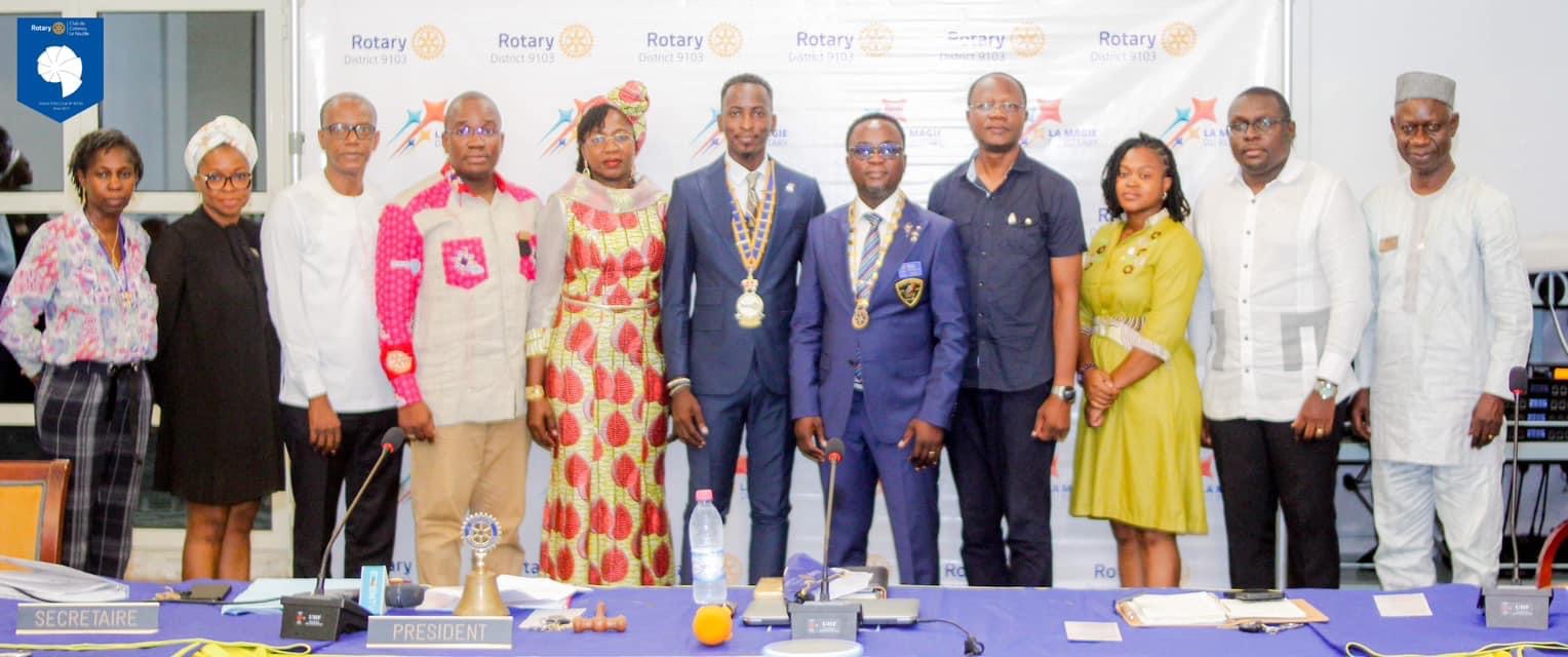 Dix personnes debout en rang devant une table avec des microphones et des documents dans une salle de réunion décorée de logos Rotary