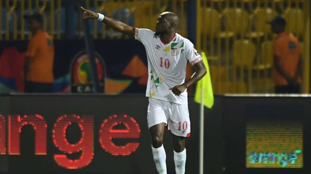 Un joueur de football en maillot blanc avec le numéro 10 lève le bras droit et pointe vers la gauche sur un terrain avec des gradins en arrière-plan