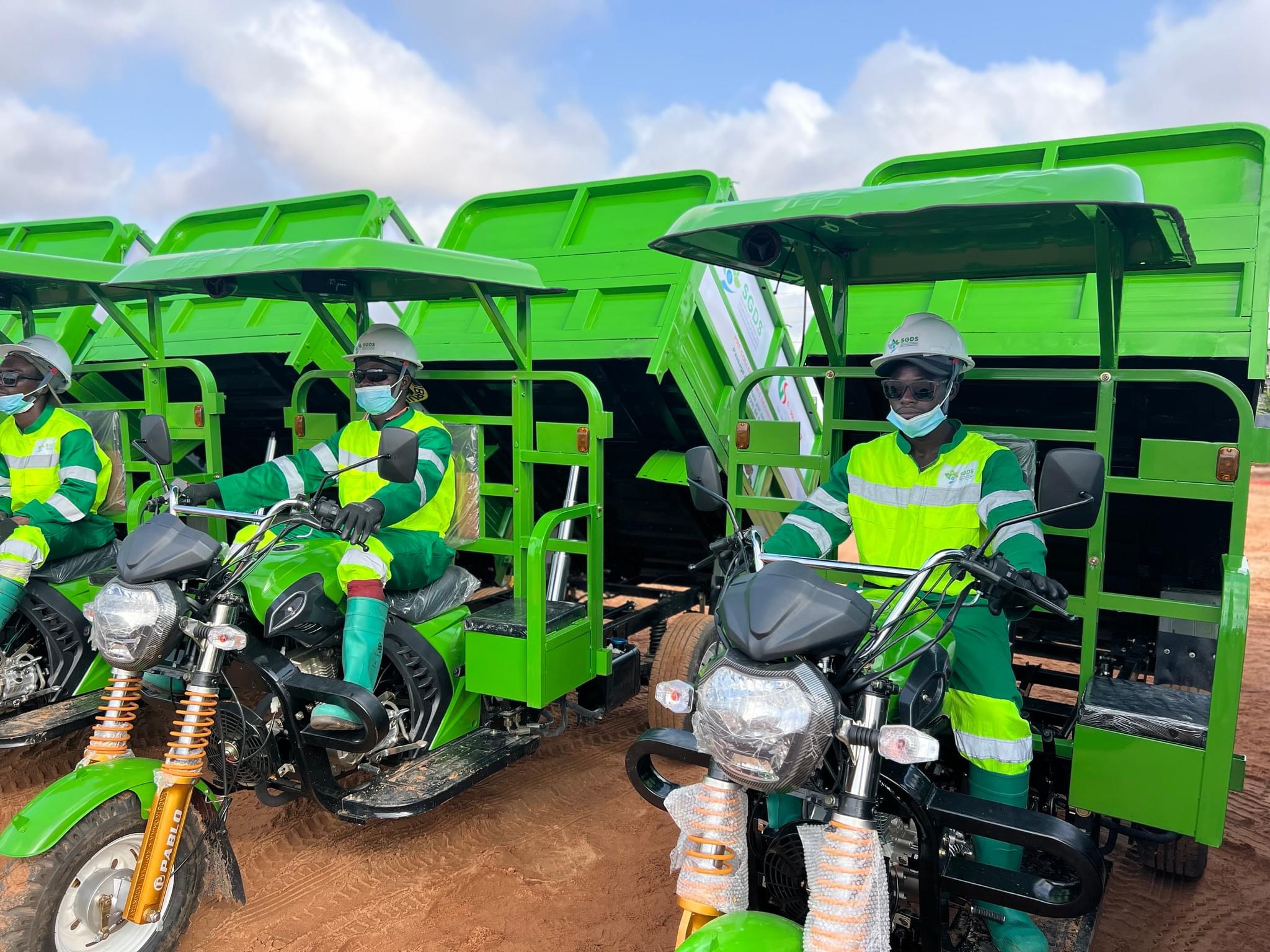 Trois hommes en tenue de protection et masques sont assis sur des motos-taxis vertes avec bennes ouvertes sur sol rougeâtre sous un ciel bleu.