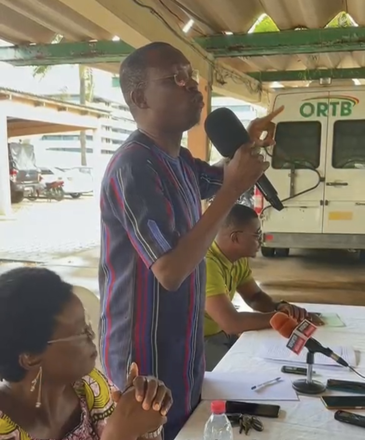 Un homme parle dans un micro debout devant une table où sont assises deux autres personnes sous un abri couvert avec un véhicule à l’arrière marqué ORTB