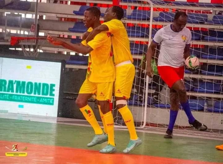 Trois joueurs en tenue de sport jouent au futsal dans un gymnase devant un but et des tribunes vides