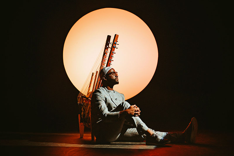 Un homme en tenue traditionnelle assis au sol devant deux instruments de musique sous un grand cercle lumineux orange