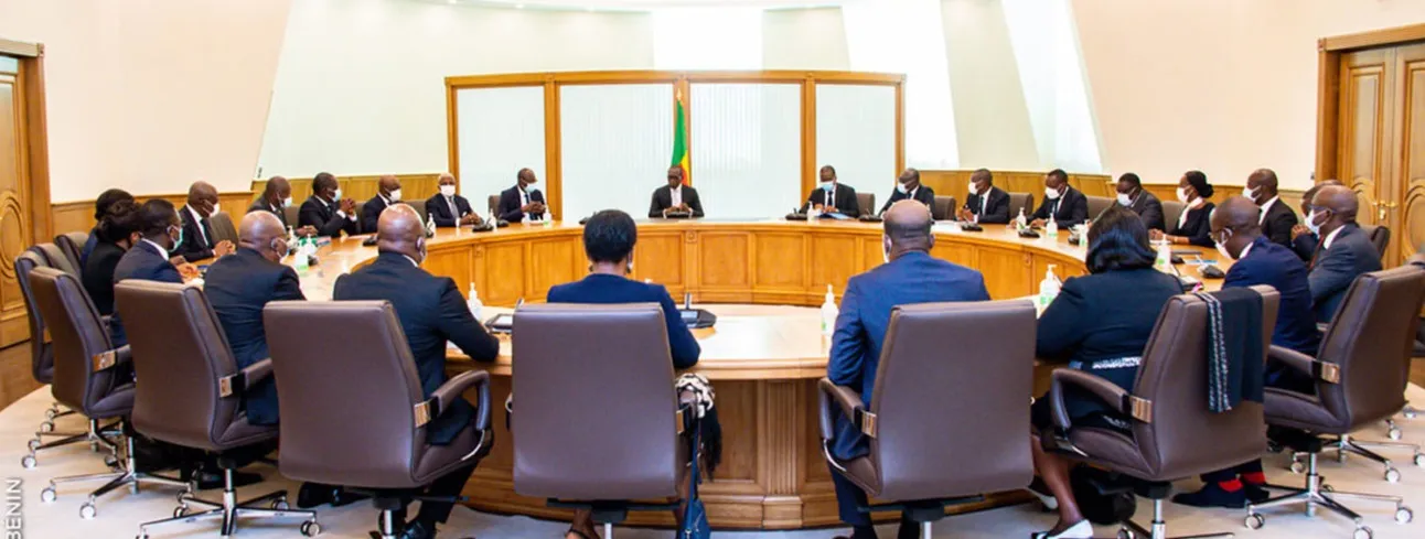 Un groupe de personnes en réunion autour d’une grande table ovale dans une salle officielle avec un drapeau derrière l’orateur central