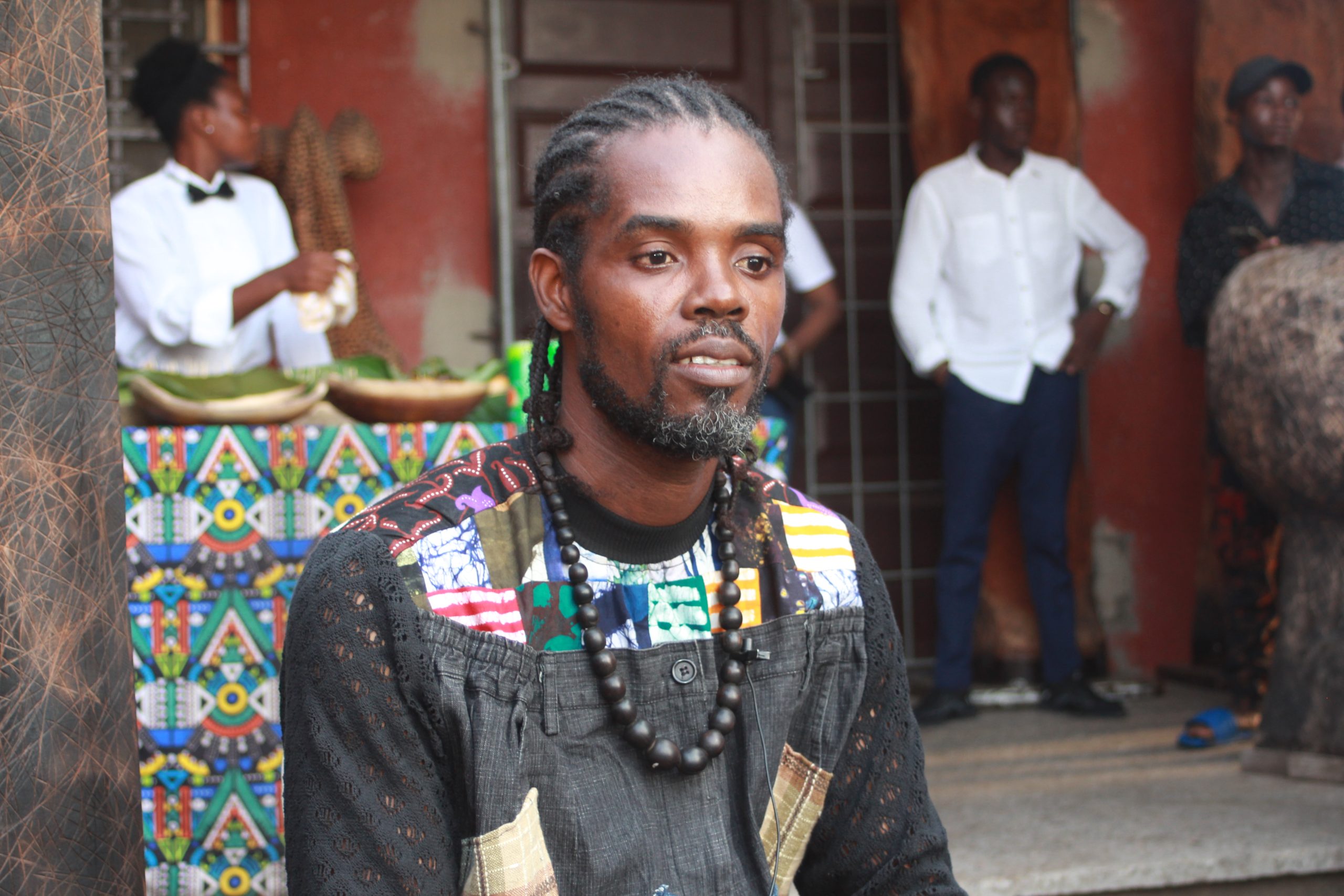 Un homme avec des dreadlocks et un collier de perles, vêtu d’un vêtement coloré, posé devant un mur rouge avec d’autres personnes floues en arrière-plan