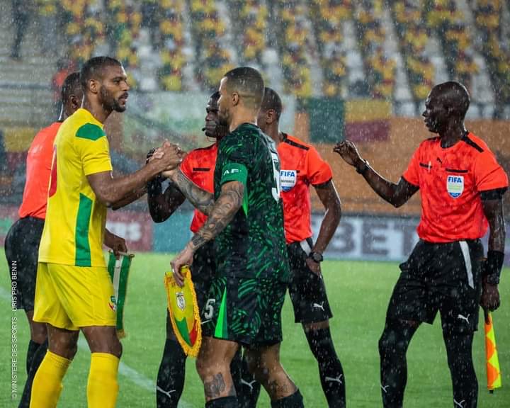 Deux joueurs de football se serrent la main sous la pluie tandis que des arbitres en tenue rouge les entourent sur le terrain