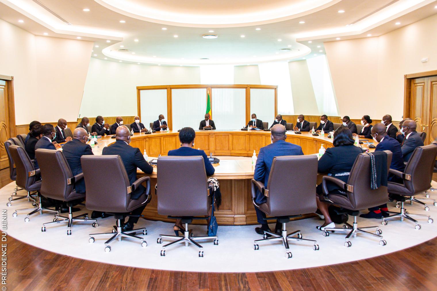 Plusieurs personnes en costume et tailleur sont assises autour d'une grande table ovale en réunion dans une salle lumineuse.