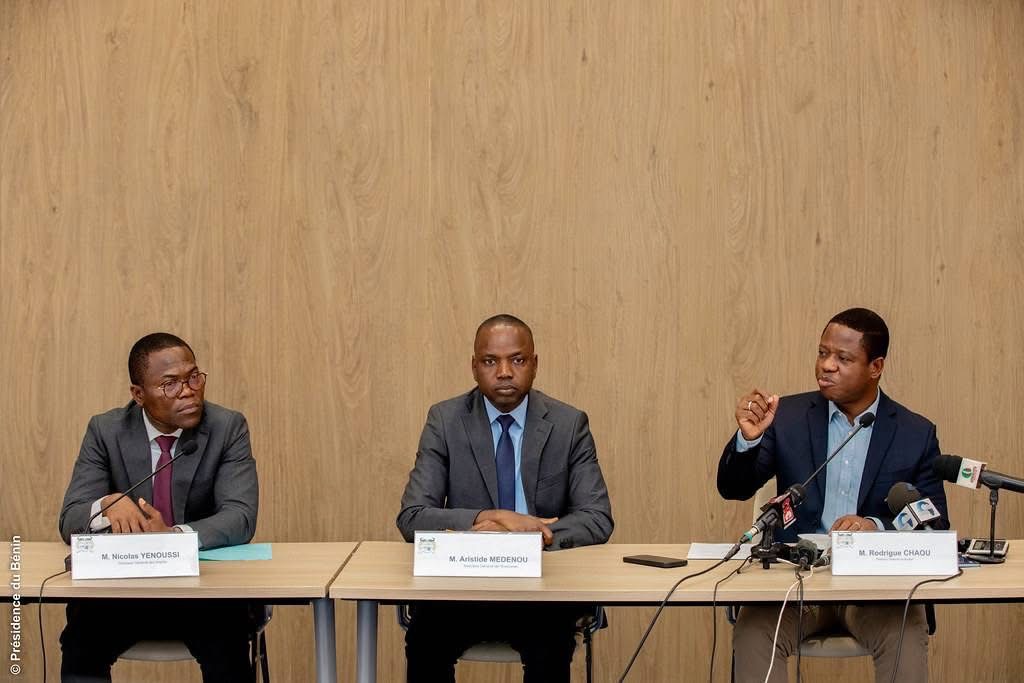 Trois hommes en costume assis derrière une table lors d'une réunion formelle avec microphones et documents posés devant eux